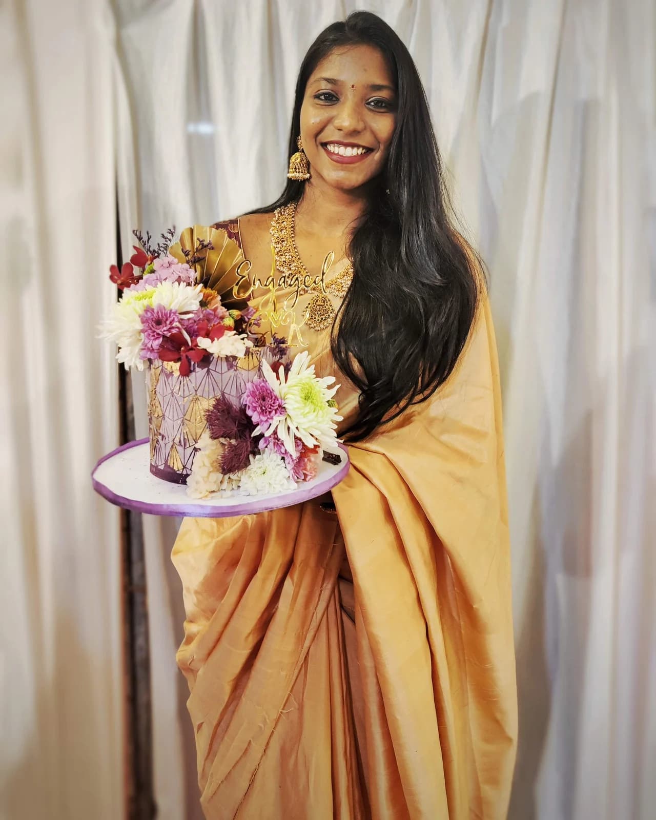 Mridula holding a purple and gold engagement cake decorated with flowers